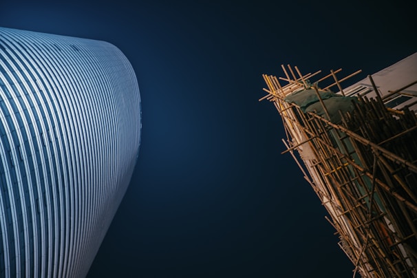 A modern skyscraper with a curved facade stands next to a building under construction covered in scaffolding. The scene is set against a deep blue evening sky, highlighting the contrast between the sleek, polished structure and the raw, unfinished construction.
