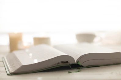 An open book with a gold-accented cover lying next to a lit candle and a cup of tea on a deep black background.