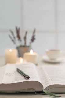 An open book resting on a wooden table beside a steaming cup of tea and a flickering candle.
