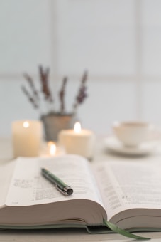 An open book lies on a table with a pen resting on its pages. In the background, several lit candles create a warm ambiance. A potted plant and a cup of tea or coffee are also visible, suggesting a cozy and peaceful setting.
