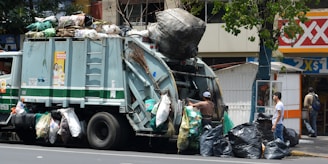 A garbage truck is parked on the street, piled high with various bags of waste. Workers are actively engaged in loading large black trash bags onto the truck. The scene is set in an urban environment with a corner convenience store visible in the background and a tree providing partial coverage above the truck.