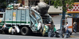 A garbage truck is parked on the street, piled high with various bags of waste. Workers are actively engaged in loading large black trash bags onto the truck. The scene is set in an urban environment with a corner convenience store visible in the background and a tree providing partial coverage above the truck.