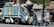 A garbage truck is parked on the street, piled high with various bags of waste. Workers are actively engaged in loading large black trash bags onto the truck. The scene is set in an urban environment with a corner convenience store visible in the background and a tree providing partial coverage above the truck.
