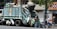 A garbage truck is parked on the street, piled high with various bags of waste. Workers are actively engaged in loading large black trash bags onto the truck. The scene is set in an urban environment with a corner convenience store visible in the background and a tree providing partial coverage above the truck.