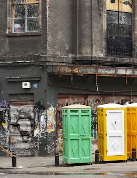 A weathered urban scene featuring a worn building facade with cracked windows, adorned with faded graffiti and rusted metal elements. Three portable toilets in green, yellow, and white stand in front of a graffitied and shuttered storefront.