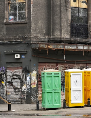 A weathered urban scene featuring a worn building facade with cracked windows, adorned with faded graffiti and rusted metal elements. Three portable toilets in green, yellow, and white stand in front of a graffitied and shuttered storefront.