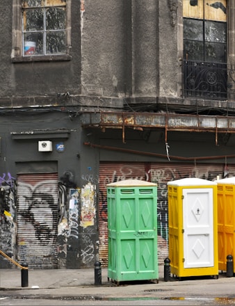 A weathered urban scene featuring a worn building facade with cracked windows, adorned with faded graffiti and rusted metal elements. Three portable toilets in green, yellow, and white stand in front of a graffitied and shuttered storefront.