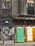 A weathered urban scene featuring a worn building facade with cracked windows, adorned with faded graffiti and rusted metal elements. Three portable toilets in green, yellow, and white stand in front of a graffitied and shuttered storefront.