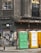 A weathered urban scene featuring a worn building facade with cracked windows, adorned with faded graffiti and rusted metal elements. Three portable toilets in green, yellow, and white stand in front of a graffitied and shuttered storefront.