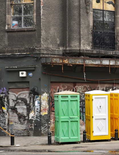 A weathered urban scene featuring a worn building facade with cracked windows, adorned with faded graffiti and rusted metal elements. Three portable toilets in green, yellow, and white stand in front of a graffitied and shuttered storefront.