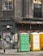 A weathered urban scene featuring a worn building facade with cracked windows, adorned with faded graffiti and rusted metal elements. Three portable toilets in green, yellow, and white stand in front of a graffitied and shuttered storefront.