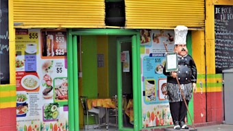 A small restaurant entrance with a colorful exterior decorated with vibrant images of food and menu prices. There's a doorway framed in green, table and chairs are visible inside. A statue of a chef holding a menu stands outside the entrance. The walls are painted in bright yellow, green, and red stripes, and there is a blackboard menu to the side listing various items.