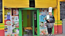 A small restaurant entrance with a colorful exterior decorated with vibrant images of food and menu prices. There's a doorway framed in green, table and chairs are visible inside. A statue of a chef holding a menu stands outside the entrance. The walls are painted in bright yellow, green, and red stripes, and there is a blackboard menu to the side listing various items.