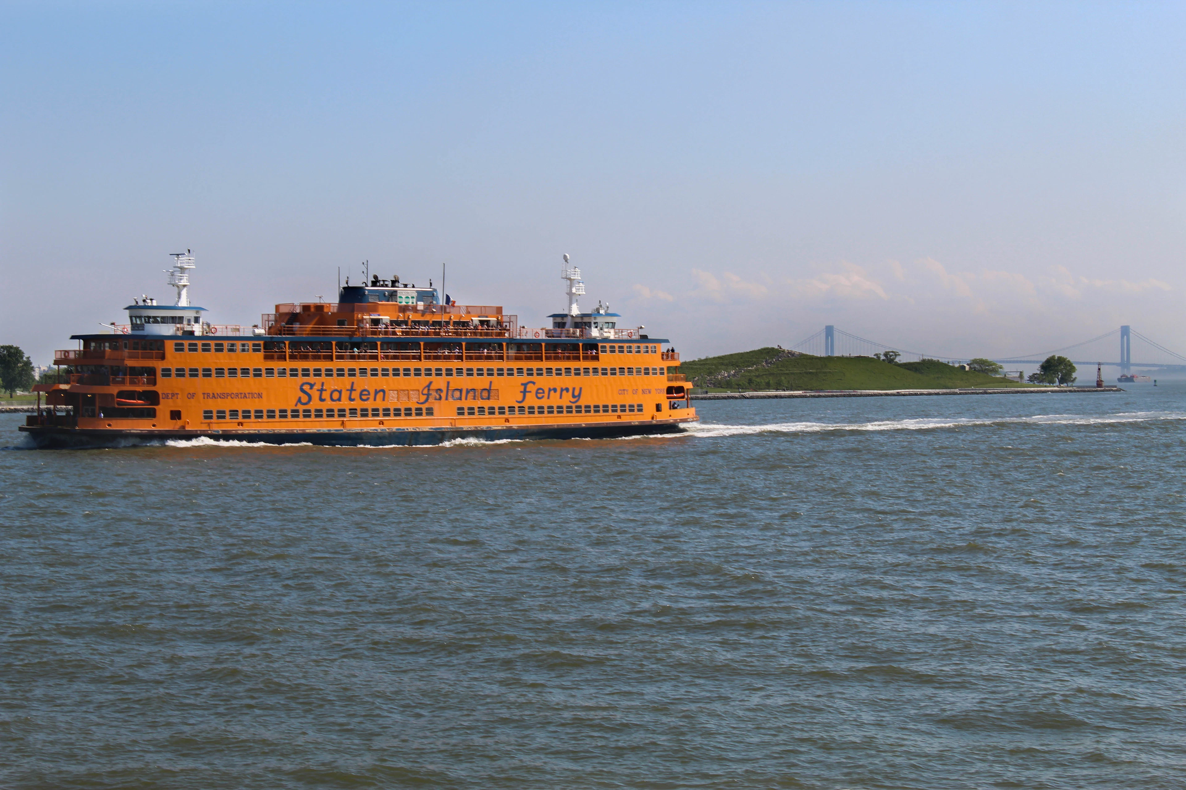 The orange Staten Island Ferry crossing New York Harbor on a bright summer day, its name painted along the hull, with the Verrazzano-Narrows Bridge spanning the water behind it