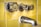 A close-up view of an old, tarnished metal bathroom faucet and knobs against a background of yellow ceramic tiles. The hardware shows signs of wear and age, with spots and discoloration visible on the metal.