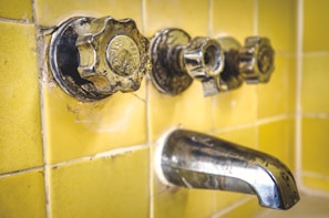 A close-up view of an old, tarnished metal bathroom faucet and knobs against a background of yellow ceramic tiles. The hardware shows signs of wear and age, with spots and discoloration visible on the metal.