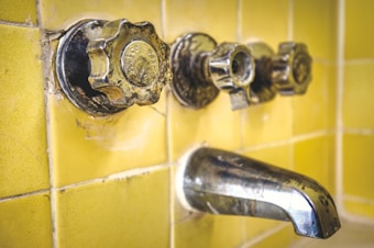 A close-up view of an old, tarnished metal bathroom faucet and knobs against a background of yellow ceramic tiles. The hardware shows signs of wear and age, with spots and discoloration visible on the metal.