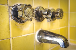 A close-up view of an old, tarnished metal bathroom faucet and knobs against a background of yellow ceramic tiles. The hardware shows signs of wear and age, with spots and discoloration visible on the metal.