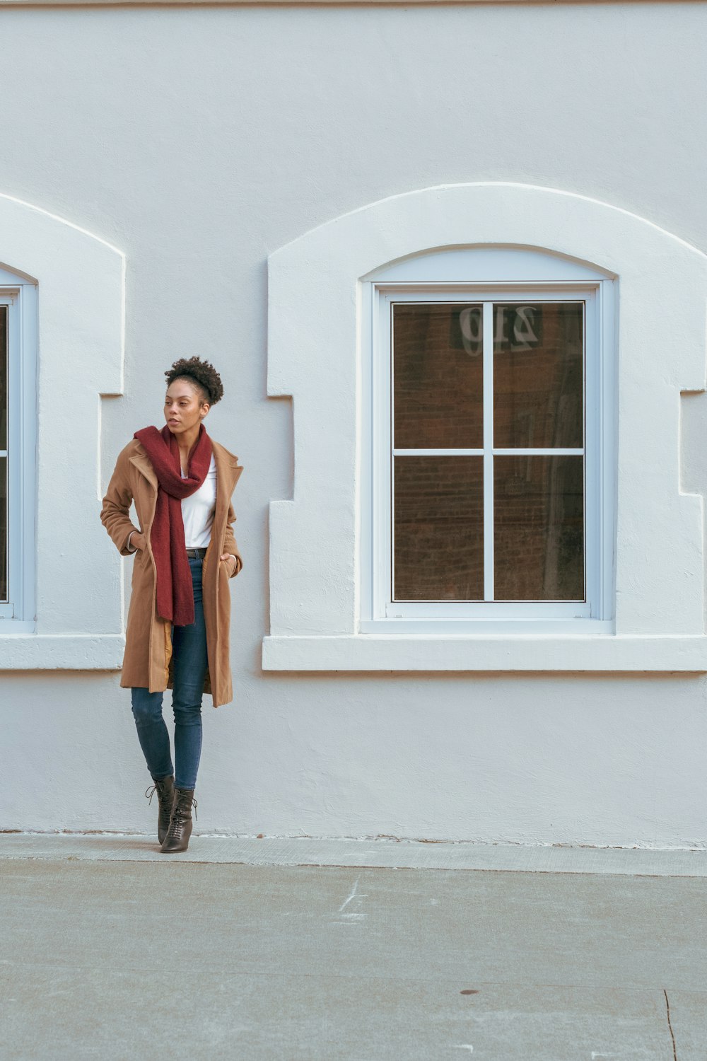 woman in brown coat and blue denim jeans standing beside white wooden window
