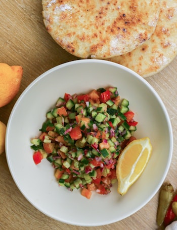 A white bowl filled with a colorful salad made of diced cucumbers, tomatoes, red onions, and possibly herbs sits on a wooden surface. Next to the salad is a lemon wedge for garnish. Surrounding the bowl are two round, golden flatbreads and some whole lemons on the table. The salad appears fresh and vibrant, and the bread looks homemade with a slightly toasted appearance.