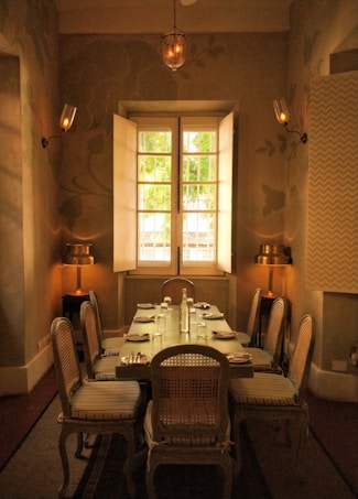 An elegantly set dining room with a wooden table surrounded by six chairs, softly lit by wall sconces and a warm hanging lamp. The room features muted tones, and natural light filters through a large window with partially opened shutters, offering a view of greenery outside.