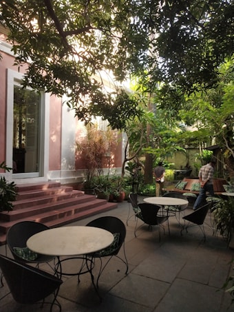 A serene outdoor seating area with several round tables and chairs surrounded by lush greenery. The space is shaded by overhanging tree branches, creating a peaceful and inviting atmosphere. There are pinkish steps leading to a building entrance, and a person can be seen walking along the garden pathway.