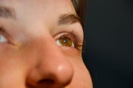 A close-up of a person's face focuses on the eye, showing intricate details of the eyelashes, eyebrow, and skin texture with a soft-focus background.