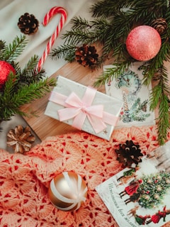 A festive arrangement includes a wrapped gift with a pink bow, nestled among evergreen branches and pinecones. A candy cane and red ornaments enhance the holiday theme, complemented by a crocheted orange fabric. A holiday card with a Christmas scene adds to the seasonal ambiance.