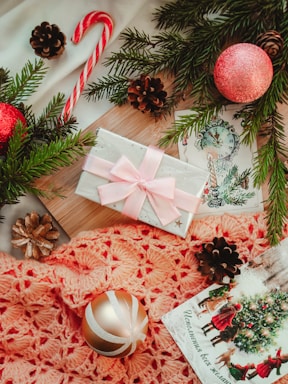 A festive arrangement includes a wrapped gift with a pink bow, nestled among evergreen branches and pinecones. A candy cane and red ornaments enhance the holiday theme, complemented by a crocheted orange fabric. A holiday card with a Christmas scene adds to the seasonal ambiance.