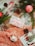 A festive arrangement includes a wrapped gift with a pink bow, nestled among evergreen branches and pinecones. A candy cane and red ornaments enhance the holiday theme, complemented by a crocheted orange fabric. A holiday card with a Christmas scene adds to the seasonal ambiance.