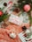 A festive arrangement includes a wrapped gift with a pink bow, nestled among evergreen branches and pinecones. A candy cane and red ornaments enhance the holiday theme, complemented by a crocheted orange fabric. A holiday card with a Christmas scene adds to the seasonal ambiance.