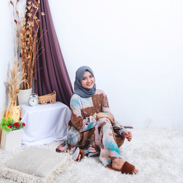 A person wearing a colorful outfit and a headscarf is sitting on a fluffy white rug. They are smiling and appear relaxed. The room has a cozy setting with a small table on the left, decorated with dried plants, a clock, and a small wicker cart. The background features a draped curtain in a muted purple shade.