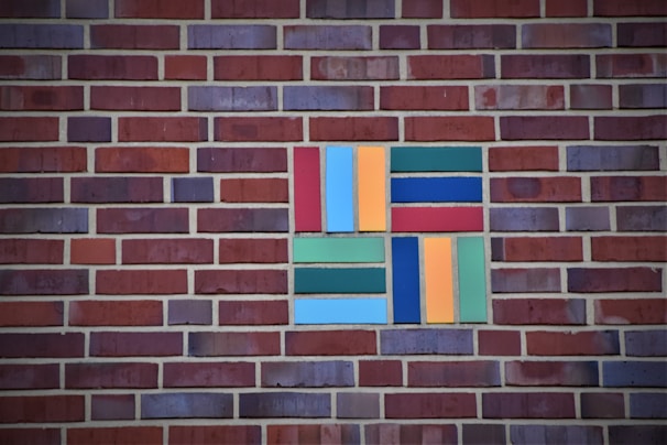 A brick wall with a patterned section of colored rectangular tiles. The tiles include shades of blue, green, orange, and red arranged in a geometric pattern.