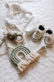 A cozy white knit sweater with small ears on the hood is laid out next to a pair of white baby sneakers with black details. A crocheted deer toy with a green scarf and wooden teething ring is placed near the sweater. Below them, a decorative woven rainbow in shades of green and off-white with tassels at the ends rests on a soft white surface.