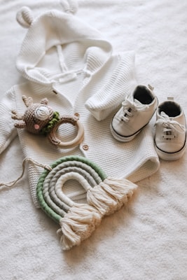 A cozy white knit sweater with small ears on the hood is laid out next to a pair of white baby sneakers with black details. A crocheted deer toy with a green scarf and wooden teething ring is placed near the sweater. Below them, a decorative woven rainbow in shades of green and off-white with tassels at the ends rests on a soft white surface.