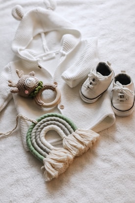 A cozy white knit sweater with small ears on the hood is laid out next to a pair of white baby sneakers with black details. A crocheted deer toy with a green scarf and wooden teething ring is placed near the sweater. Below them, a decorative woven rainbow in shades of green and off-white with tassels at the ends rests on a soft white surface.