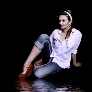 A woman with a retro style sits on an unseen surface with water reflection effects. She wears a white blouse, rolled-up jeans, and red heels. Her hair is styled with a polka dot headband, and she wears statement earrings. A tattoo is visible on her ankle.