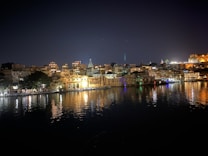 A nighttime cityscape with well-lit buildings reflecting on the calm, dark water of a lake. The skyline is adorned with architectural structures, some of which are ornately illuminated. The soft glow of lights creates a serene and tranquil atmosphere.
