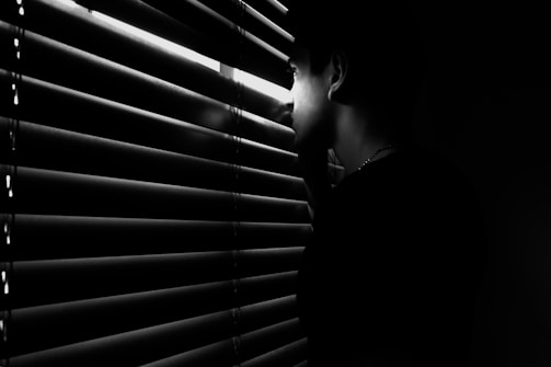 A person is peering through partially opened blinds in a dimly lit setting. The shadows cast by the blinds create a striped pattern across their face. The overall mood is mysterious and contemplative.
