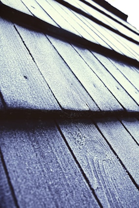 Close-up view of textured roof shingles covered in frost or ice, with a diagonal perspective that draws the eye across the pattern. The surface appears rough, with a light dusting of frost giving a sparkling effect under the light.