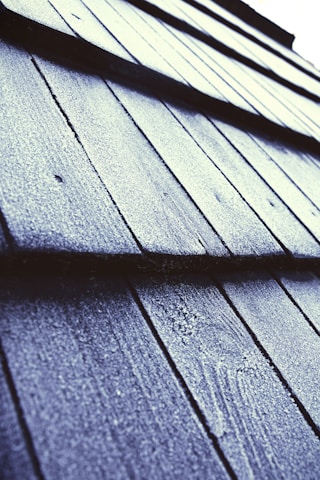 Close-up view of textured roof shingles covered in frost or ice, with a diagonal perspective that draws the eye across the pattern. The surface appears rough, with a light dusting of frost giving a sparkling effect under the light.