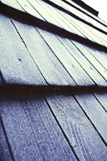 Close-up view of textured roof shingles covered in frost or ice, with a diagonal perspective that draws the eye across the pattern. The surface appears rough, with a light dusting of frost giving a sparkling effect under the light.