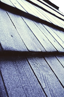 Close-up view of textured roof shingles covered in frost or ice, with a diagonal perspective that draws the eye across the pattern. The surface appears rough, with a light dusting of frost giving a sparkling effect under the light.