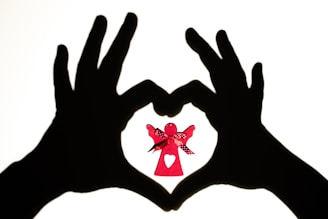 Two hands are silhouetted against a light background, forming a heart shape. Inside this heart shape is a small red paper angel with a decorative ribbon and a heart cut-out on its dress.