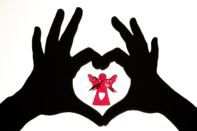 Two hands are silhouetted against a light background, forming a heart shape. Inside this heart shape is a small red paper angel with a decorative ribbon and a heart cut-out on its dress.