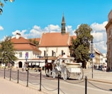 A picturesque street scene featuring a horse-drawn carriage and a small tour vehicle in front of historic European-style buildings. The buildings have red-tiled roofs and are surrounded by lush green trees under a bright blue sky with scattered clouds.
