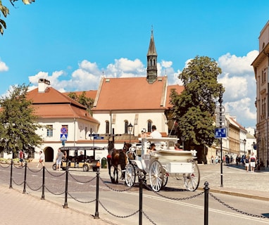 A picturesque street scene featuring a horse-drawn carriage and a small tour vehicle in front of historic European-style buildings. The buildings have red-tiled roofs and are surrounded by lush green trees under a bright blue sky with scattered clouds.