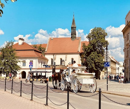 A picturesque street scene featuring a horse-drawn carriage and a small tour vehicle in front of historic European-style buildings. The buildings have red-tiled roofs and are surrounded by lush green trees under a bright blue sky with scattered clouds.