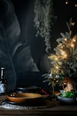 A cozy, dimly lit dining scene with a small decorative Christmas tree adorned with warm white lights. The table is set with a glass of wine, a bottle, and a plate, creating an intimate holiday atmosphere. Hanging greenery and dark, textured background enhance the festive setting.