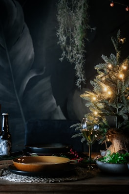 A cozy, dimly lit dining scene with a small decorative Christmas tree adorned with warm white lights. The table is set with a glass of wine, a bottle, and a plate, creating an intimate holiday atmosphere. Hanging greenery and dark, textured background enhance the festive setting.